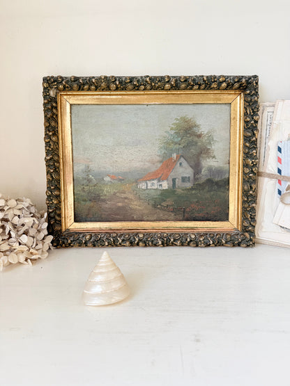 A framed vintage landscape painting of a small white house with a red roof, displayed on a white surface with decorative props.