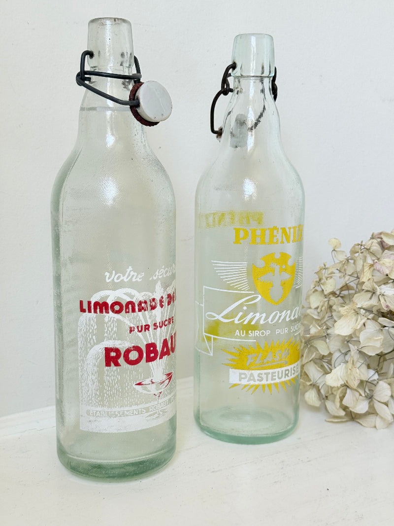 Pair of vintage French glass bottles with retro typography and graphics, photographed on a white surface