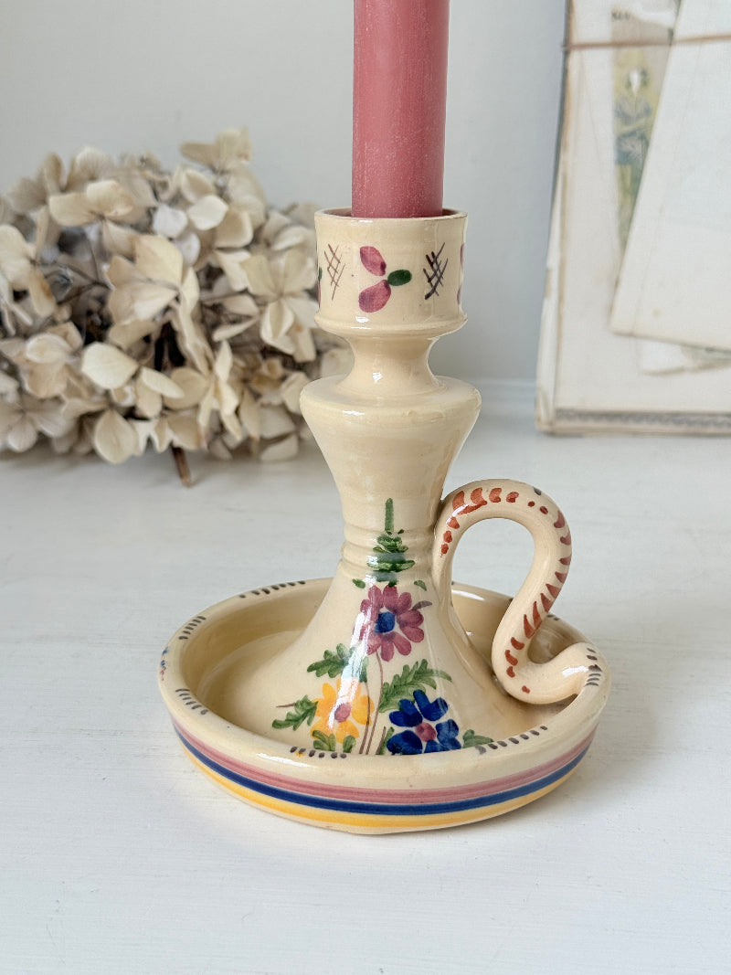 A cream-colored ceramic candlestick holder with hand-painted floral motifs and a snake handle, holding a pink candle, set against a softly blurred background of dried flowers and framed art.