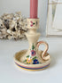 A cream-colored ceramic candlestick holder with hand-painted floral motifs and a snake handle, holding a pink candle, set against a softly blurred background of dried flowers and framed art.