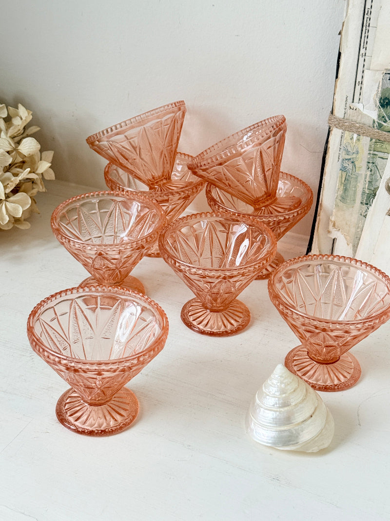 Six pink cut-glass cocktail coupe glasses arranged on a white surface with a white shell and dried flowers as props.