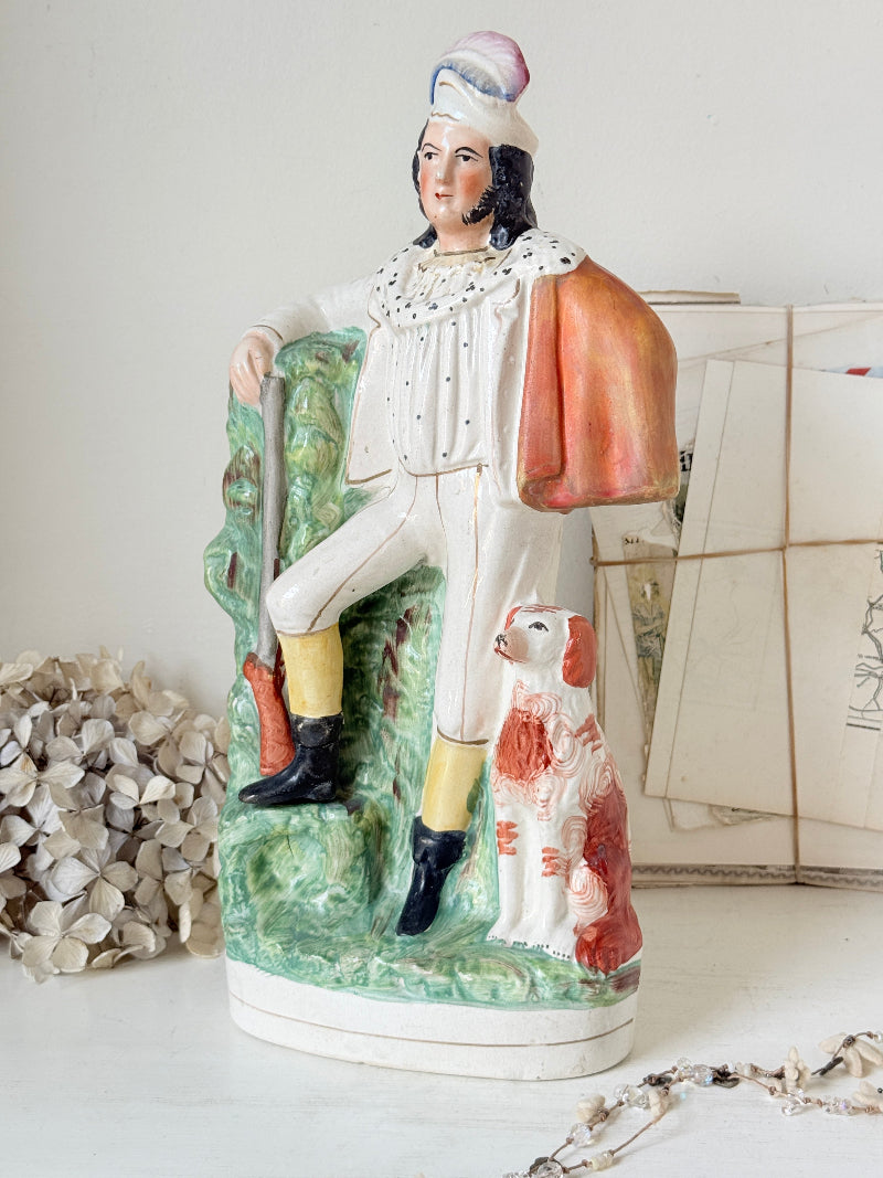 A vintage hand-painted ceramic figurine of a man in historical white and yellow attire with a pink hat, seated on a green landscape base next to a white and red dog, displayed on a tabletop with antique papers in the background.