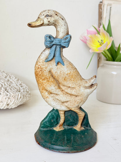 Vintage-style duck figurine with a speckled cream body, blue bow, and green base, displayed on a tabletop with soft home decor props in the background.