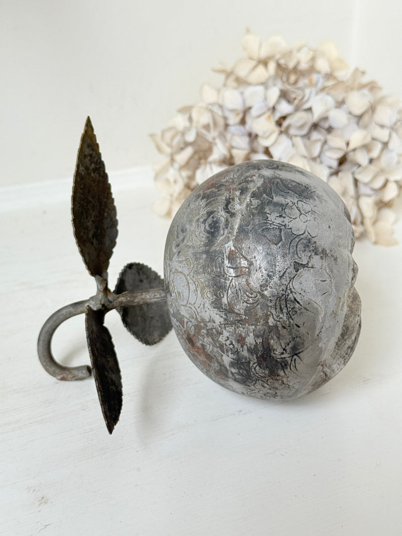 A decorative vintage-style metal doorbell with a round textured skull face and a leaf-shaped pull, displayed on a white surface with dried flowers in the background.