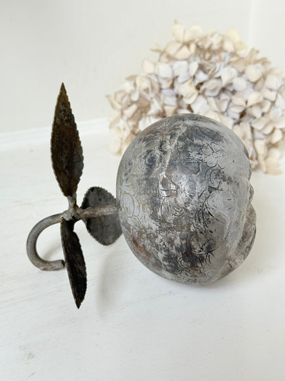 A decorative vintage-style metal doorbell with a round textured skull face and a leaf-shaped pull, displayed on a white surface with dried flowers in the background.