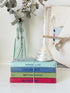A stack of four decorative books tied with twine sits on a white surface, styled with a glass bottle vase holding dried leaves, a small white shell, and a folded cream fabric item in the background.