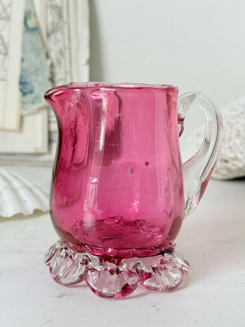 Antique cranberry glass cream jug with clear applied handle, crimped pedestal foot and deep pink hand blown glass body
