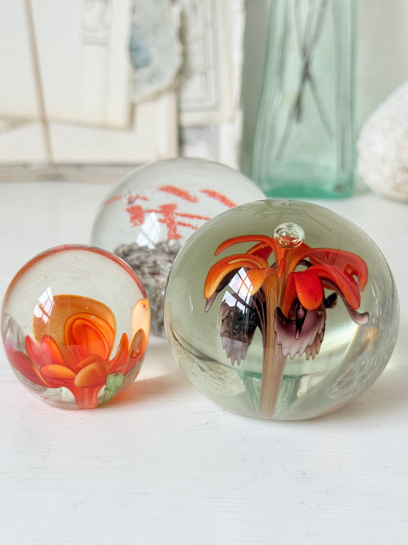 Trio of orange flower glass paperweights with green stems and controlled bubbles, photographed on a white surface
