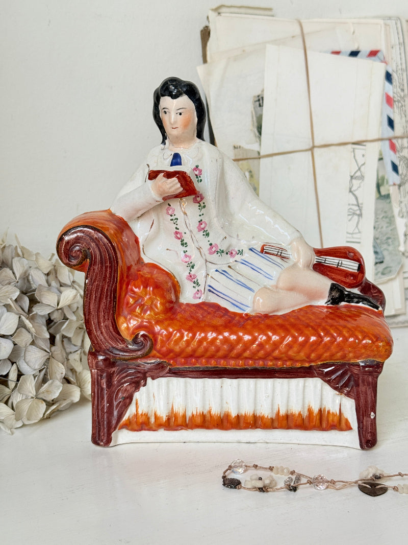 A vintage ceramic figurine of a man reclining on an orange-red chaise lounge and reading, displayed on a white surface with decorative dried flowers and old envelopes in the background.