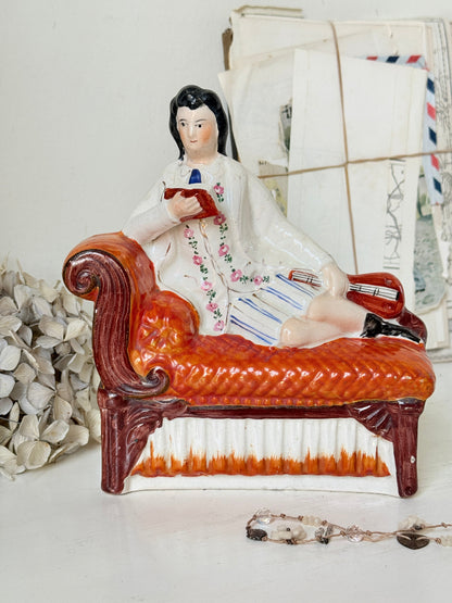 A vintage ceramic figurine of a man reclining on an orange-red chaise lounge and reading, displayed on a white surface with decorative dried flowers and old envelopes in the background.