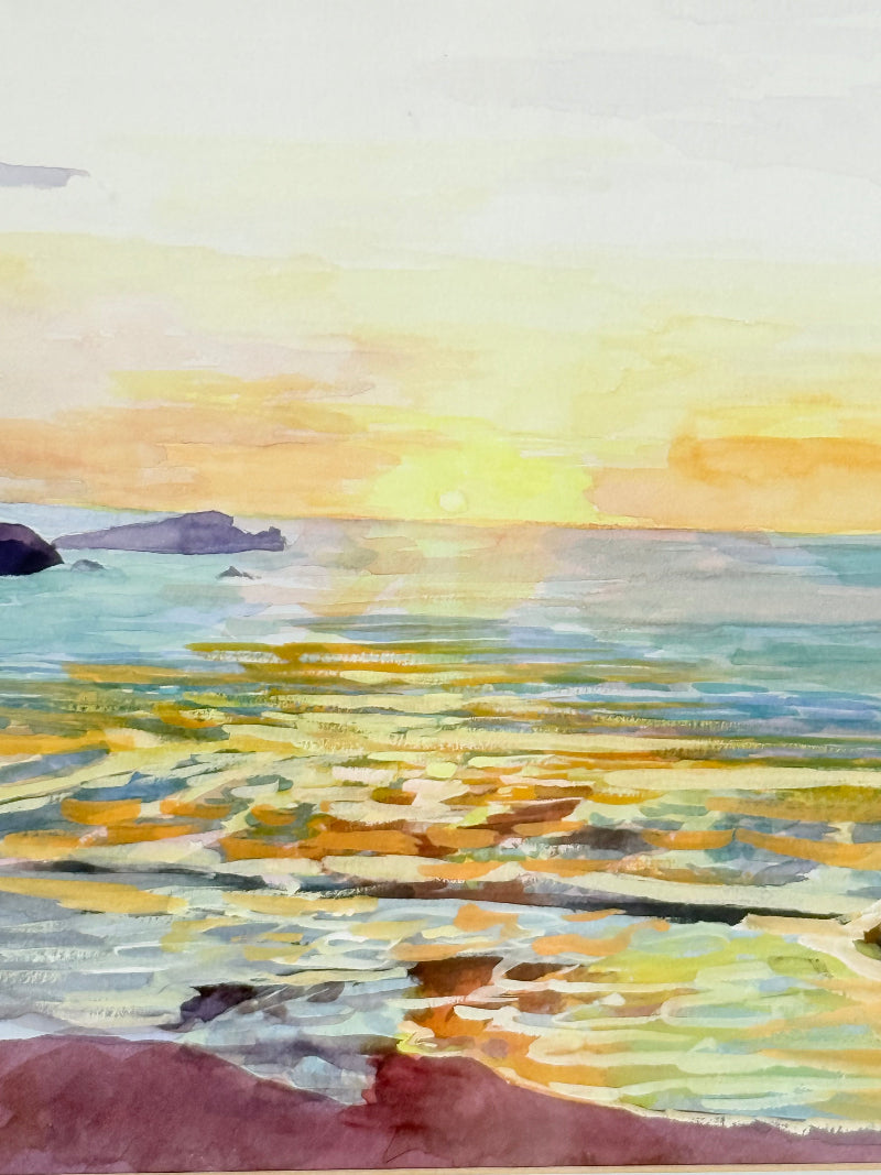 Framed coastal watercolour of sunset over sea with rocks in foreground, in gilt frame with pale mount, photographed on white surface.