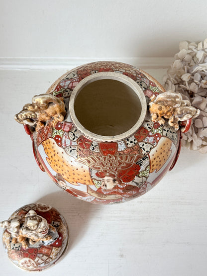 An ornate ceramic ginger jar with a matching lid, decorated with detailed hand-painted figures and animals in red, gold, and white, set against a white background.
