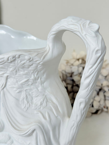 A close-up of a white ceramic pitcher with intricate embossed floral and leaf patterns, featuring a curved handle and a soft-focus decorative background.