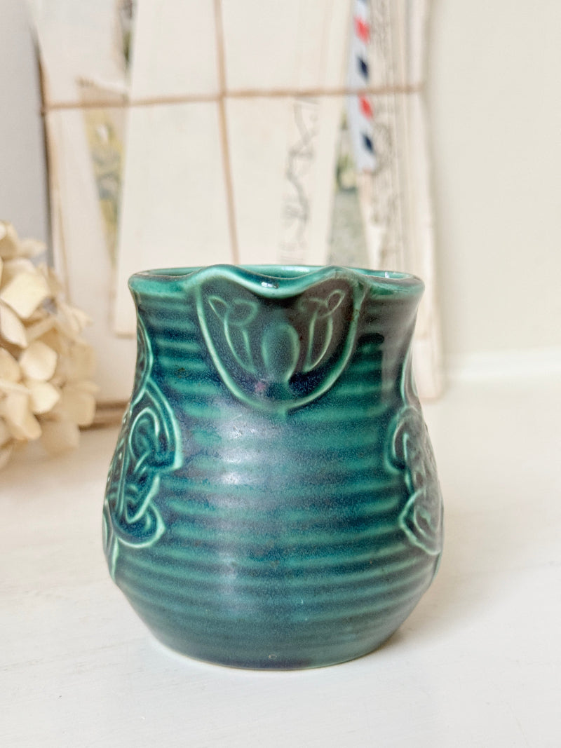 A small teal-green ceramic pitcher with a ribbed body, decorative embossed floral motifs, and a flared rim, photographed on a light surface with a softly blurred background.
