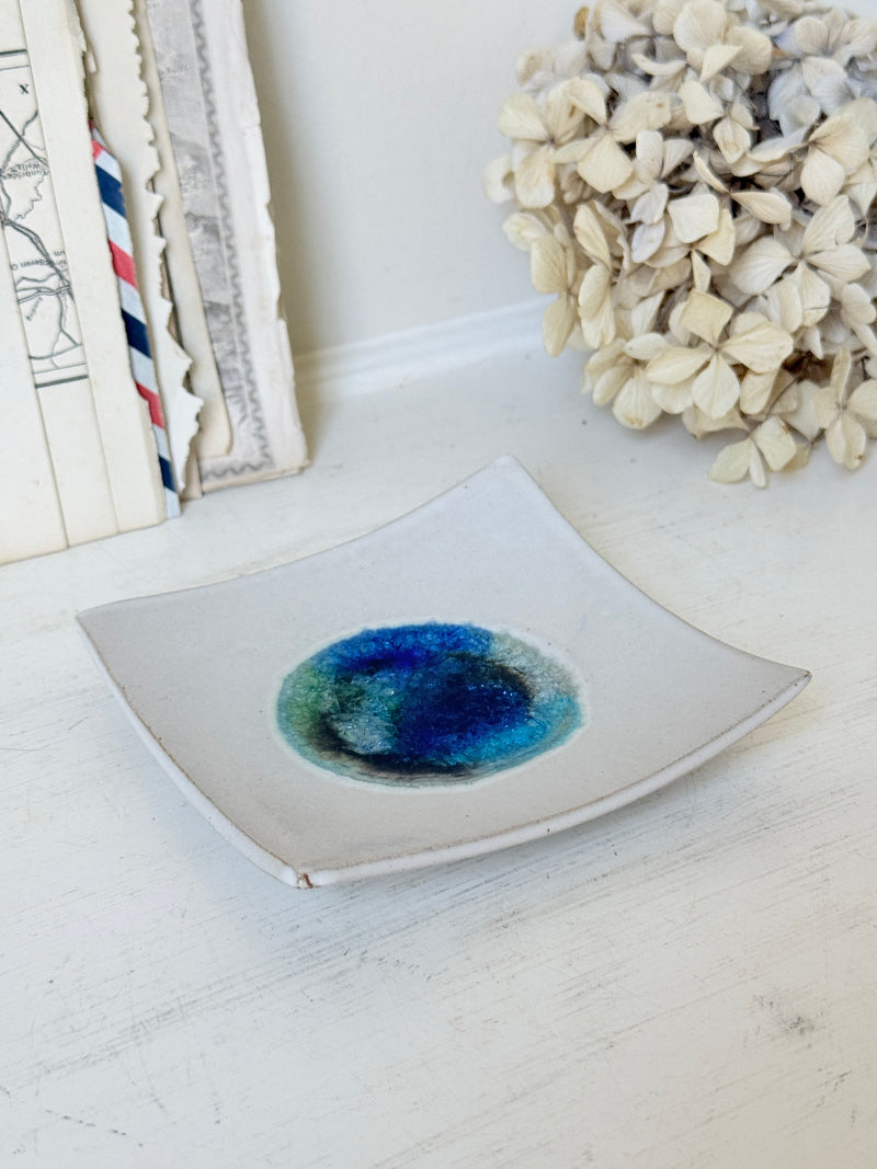 A square white ceramic dish with a curved bottom edge, featuring a hand-painted blue and green watercolor design in the center, styled on a white wooden surface with dried flowers and books in the background.