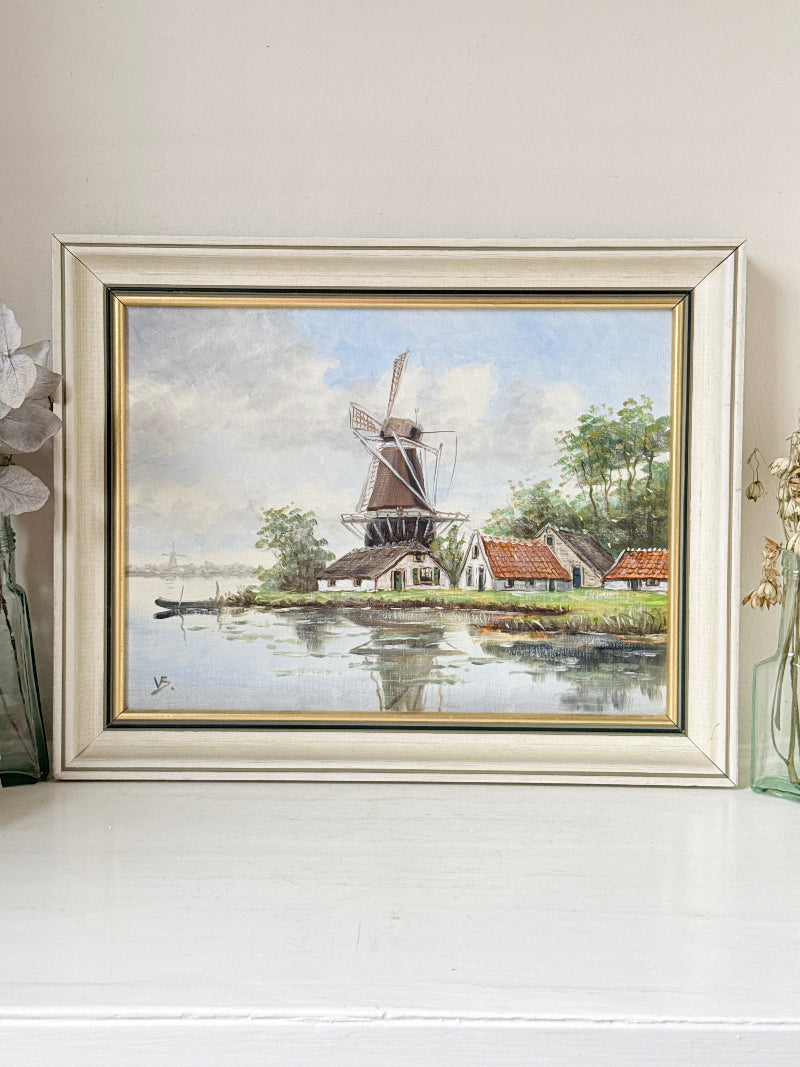 A framed landscape painting of a Dutch windmill by a canal, displayed on a white shelf against a beige wall. The frame is a distressed light beige with gold inner trim.