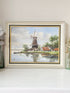 A framed landscape painting of a Dutch windmill by a canal, displayed on a white shelf against a beige wall. The frame is a distressed light beige with gold inner trim.
