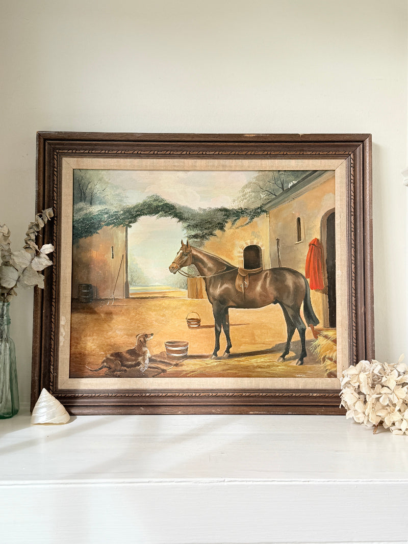 A framed vintage oil painting depicting a brown horse and dog in a stable, displayed on a white mantelpiece with decorative dried flowers and a shell.