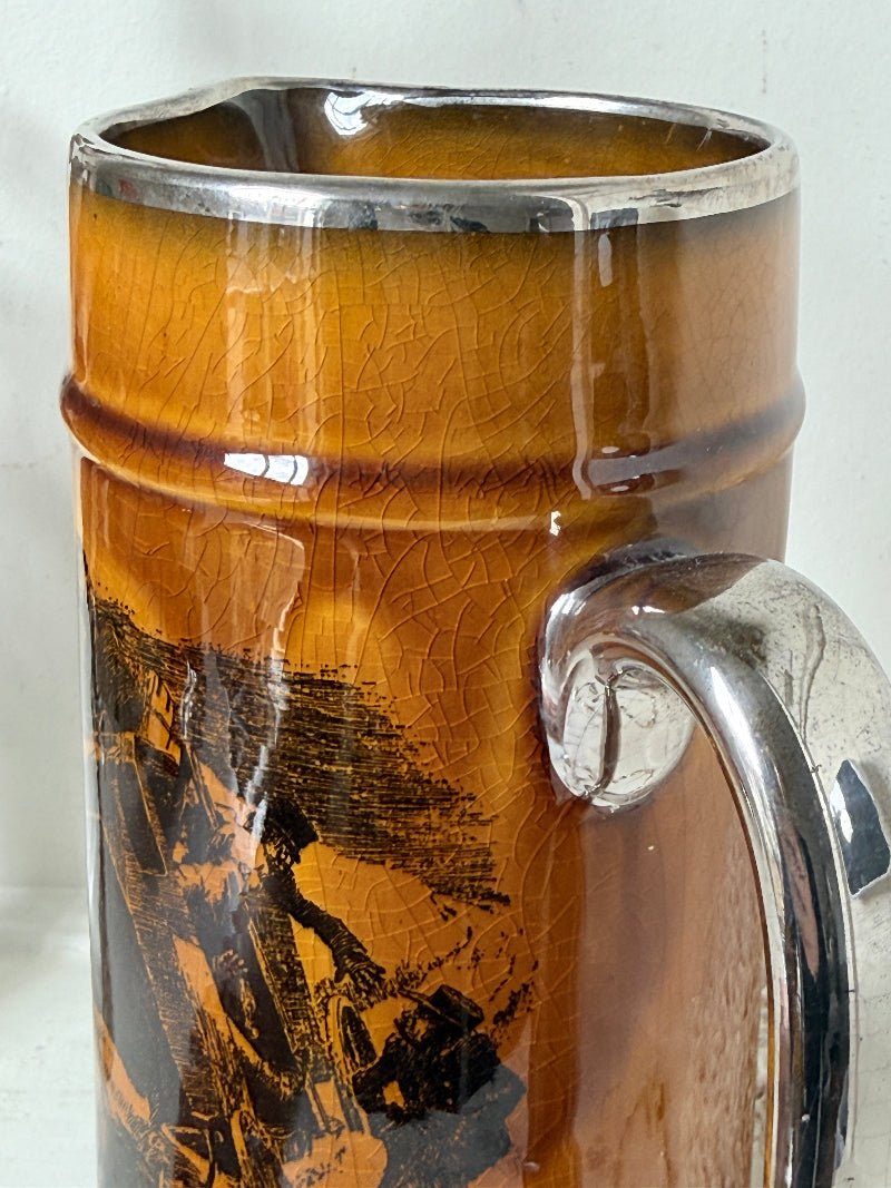 Antique Ridgways stoneware tall jug photographed on a white surface in amber treacle glaze with black transfer scenes and silver tone rim