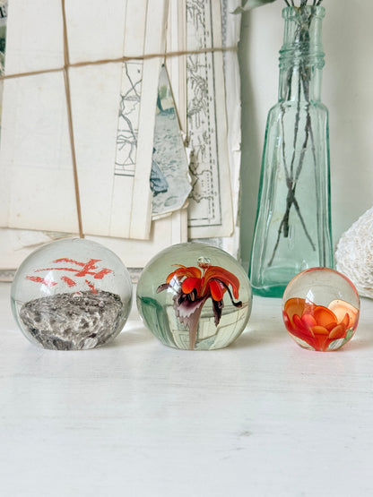 Trio of orange flower glass paperweights with green stems and controlled bubbles, photographed on a white surface