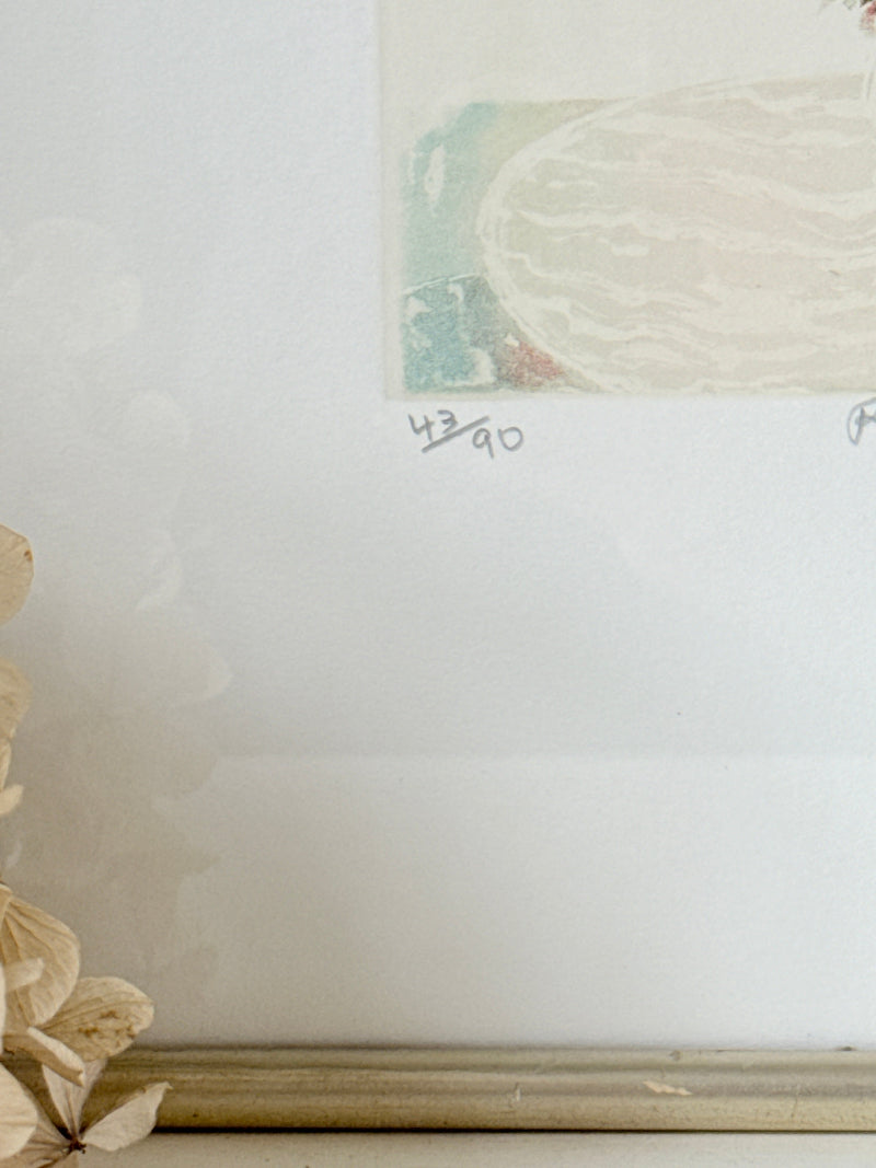 A close-up of a framed art print with a white mat and a gold-toned frame, showing part of a pastel illustration and handwritten edition markings &