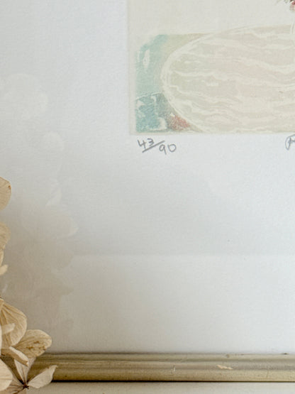 A close-up of a framed art print with a white mat and a gold-toned frame, showing part of a pastel illustration and handwritten edition markings &