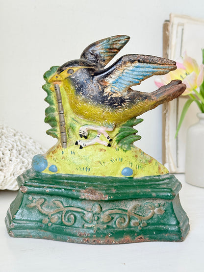 A vintage hand-painted cast iron bird feeder and money box featuring a robin holding a penny, set against a softly blurred background with a white vase and decorative items.
