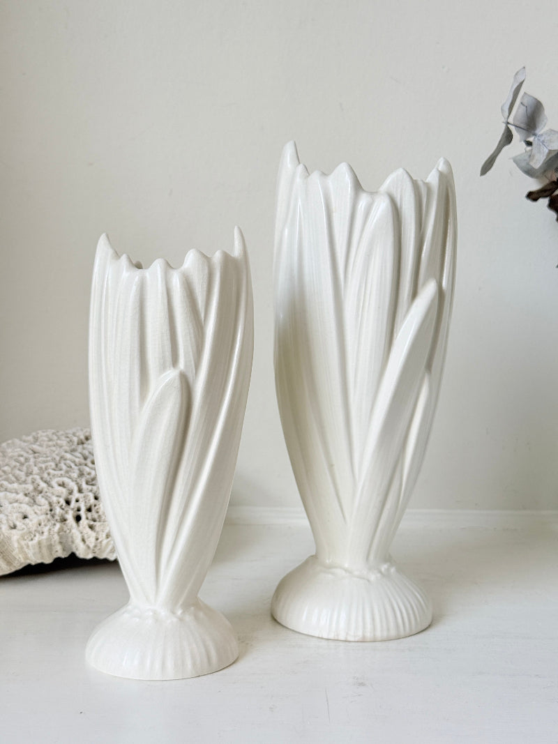 Two white ceramic tulip-shaped vases with fluted bases, displayed on a white surface against a light beige wall.