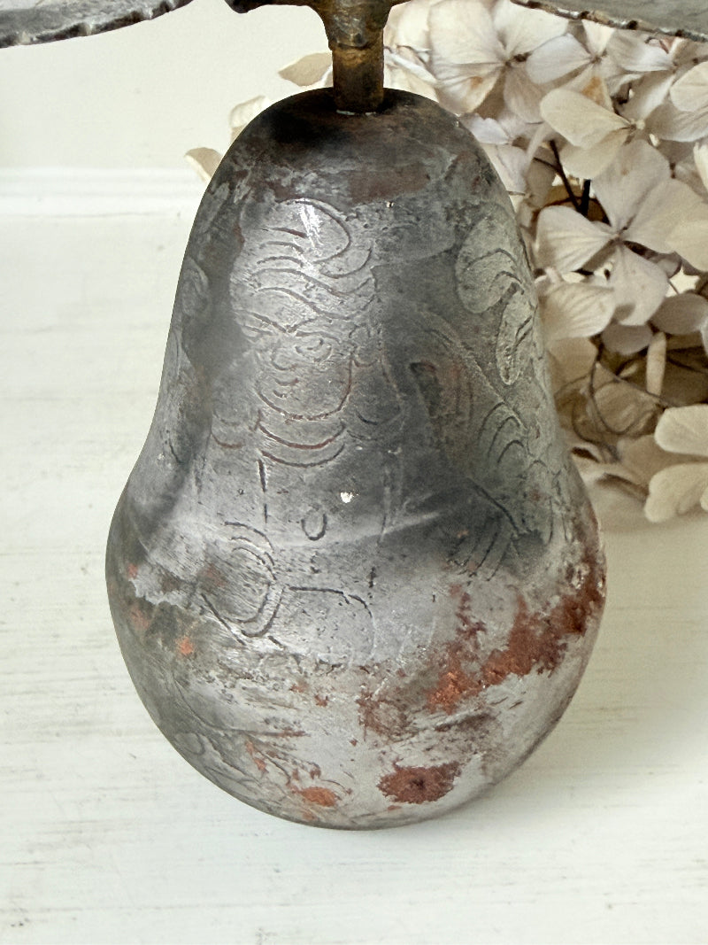 A vintage distressed metal pear-shaped object with embossed floral patterns, resting on a white wooden surface next to dried white flowers.