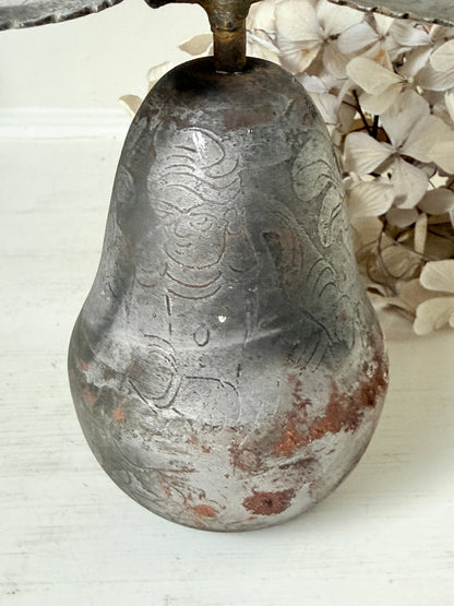 A vintage distressed metal pear-shaped object with embossed floral patterns, resting on a white wooden surface next to dried white flowers.