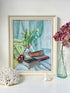 A framed still life painting displayed on a white surface against a white wall, featuring a vase of yellow flowers, stacked books, and red apples. The frame is light wood, and the scene is styled with a coral shell, a small seashell, and a green glass bottle with dried pink flowers.