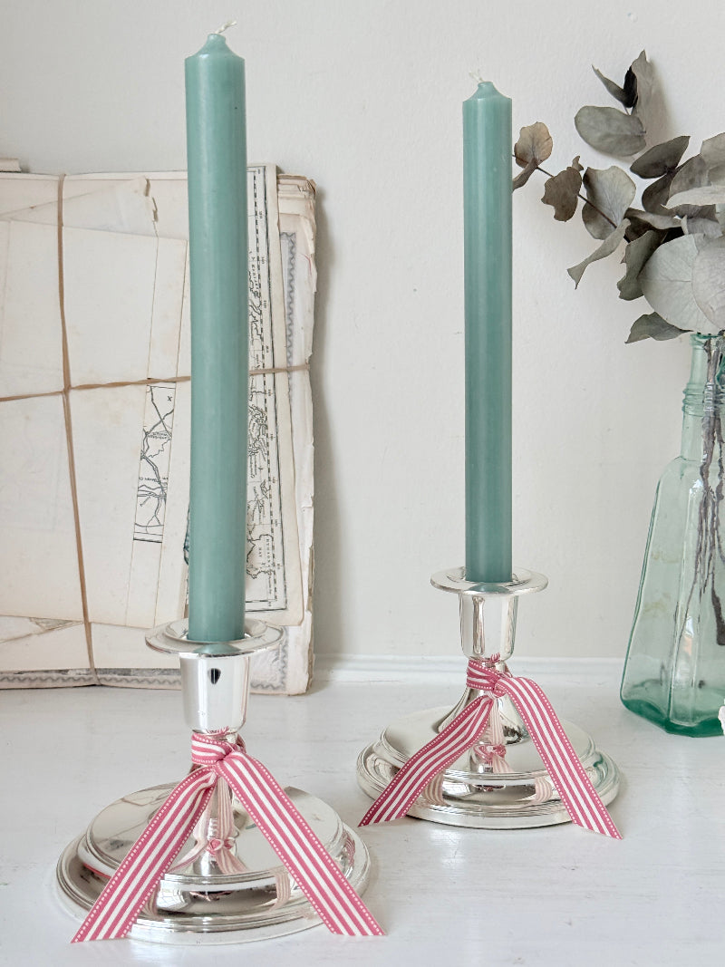 Pair of silver tone candlesticks with wide round stepped bases holding green candles, photographed on a white surface