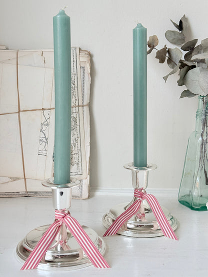 Pair of silver tone candlesticks with wide round stepped bases holding green candles, photographed on a white surface