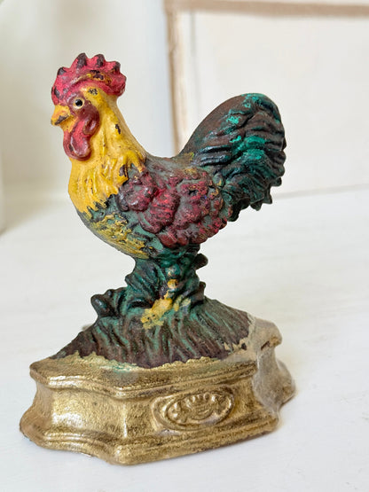 A vintage cast iron rooster figurine standing on a decorative base, featuring a yellow head and chest, red comb and wattle, and a dark green and red tail, set against a softly blurred light background.