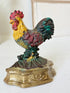 A vintage cast iron rooster figurine standing on a decorative base, featuring a yellow head and chest, red comb and wattle, and a dark green and red tail, set against a softly blurred light background.
