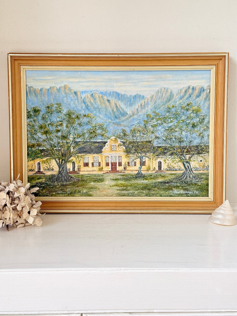 A framed landscape oil painting depicting a yellow colonial-style building surrounded by green trees, set against blue mountains under a light sky. The painting is housed in a wide, light brown wooden frame and displayed on a white mantelpiece or shelf next to dried flowers and a small shell.