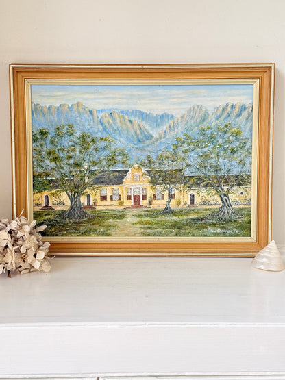 A framed landscape oil painting depicting a yellow colonial-style building surrounded by green trees, set against blue mountains under a light sky. The painting is housed in a wide, light brown wooden frame and displayed on a white mantelpiece or shelf next to dried flowers and a small shell.