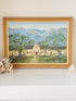 A framed landscape oil painting depicting a yellow colonial-style building surrounded by green trees, set against blue mountains under a light sky. The painting is housed in a wide, light brown wooden frame and displayed on a white mantelpiece or shelf next to dried flowers and a small shell.