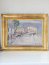 A framed impressionist landscape painting depicting a street scene with buildings, a tree, and figures, housed in a wide ornate gold frame and resting on a white surface against a white wall.