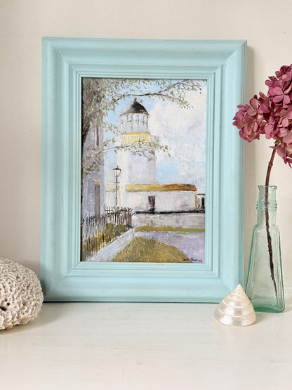 A framed art print depicting a lighthouse scene, styled on a white surface with a glass vase of pink hydrangeas, a white shell, and a textured white object against a beige wall.