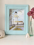 A framed art print depicting a lighthouse scene, styled on a white surface with a glass vase of pink hydrangeas, a white shell, and a textured white object against a beige wall.