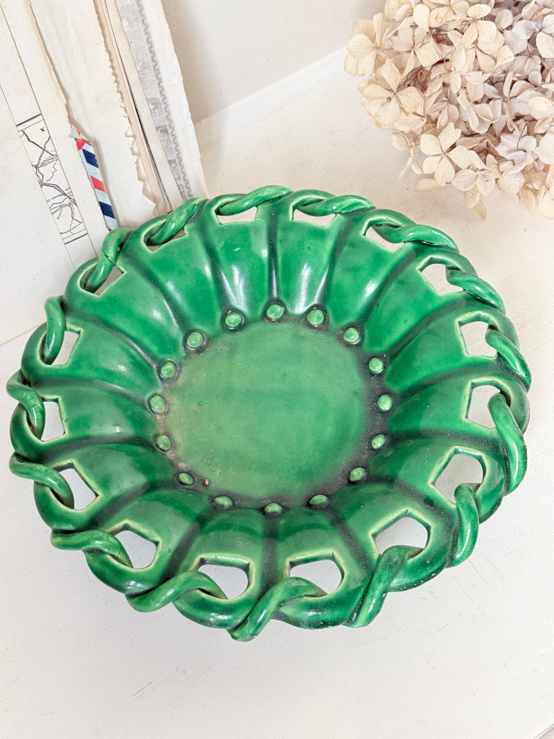 A green ceramic decorative bowl with a scalloped, openwork edge, displayed on a light surface with neutral decor in the background.