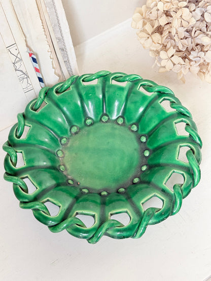 A green ceramic decorative bowl with a scalloped, openwork edge, displayed on a light surface with neutral decor in the background.