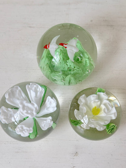 Trio of white flower glass paperweights in graduated sizes with green leaves, photographed on a white surface