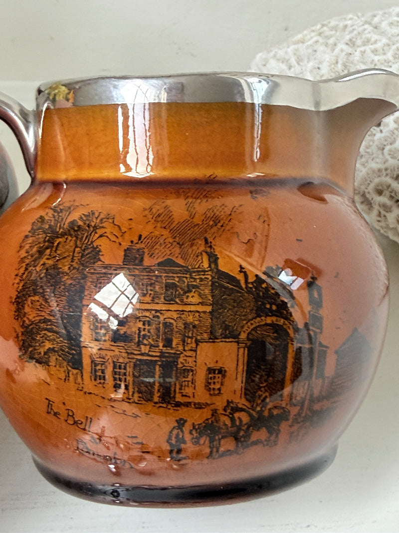 Pair of antique Ridgways stoneware creamer jugs photographed on a white surface in amber treacle glaze with black coaching scenes and silver lustre rims