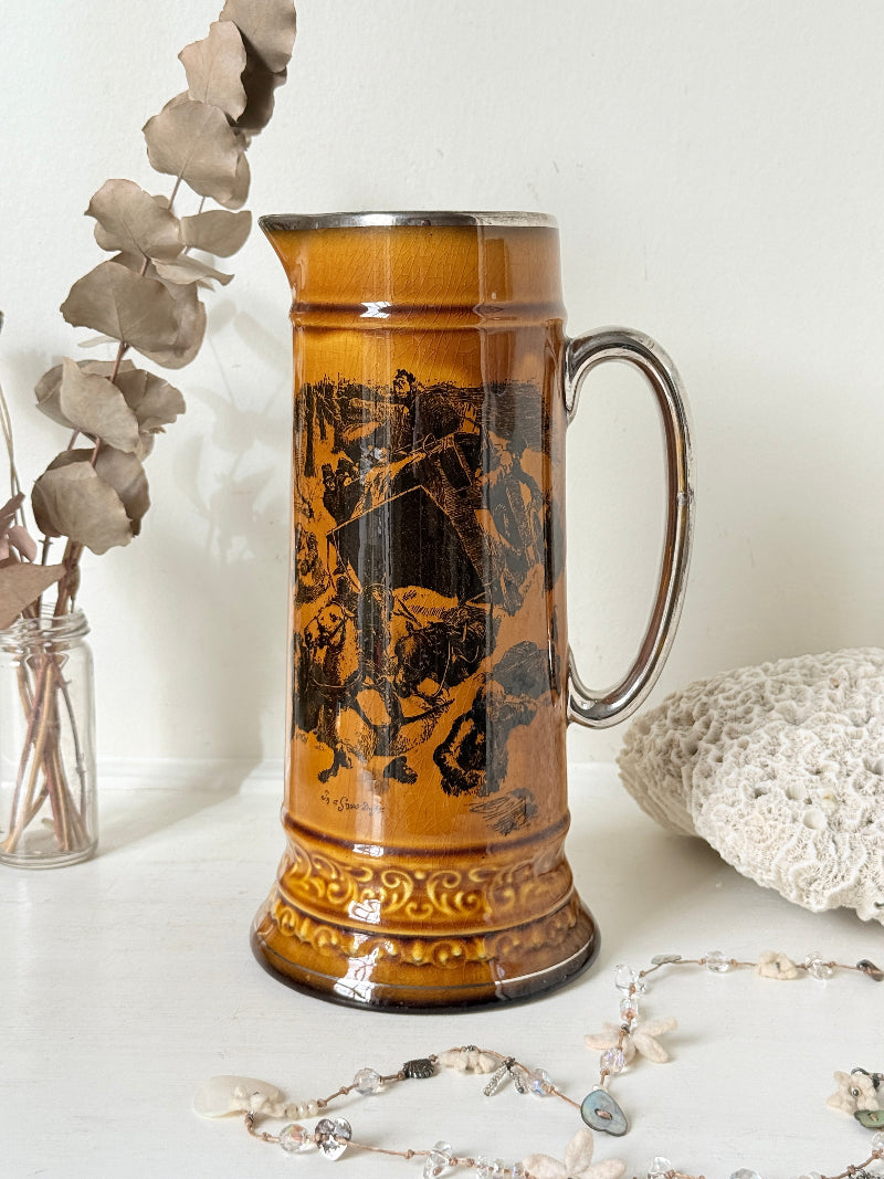 Antique Ridgways stoneware tall jug photographed on a white surface in amber treacle glaze with black transfer scenes and silver tone rim