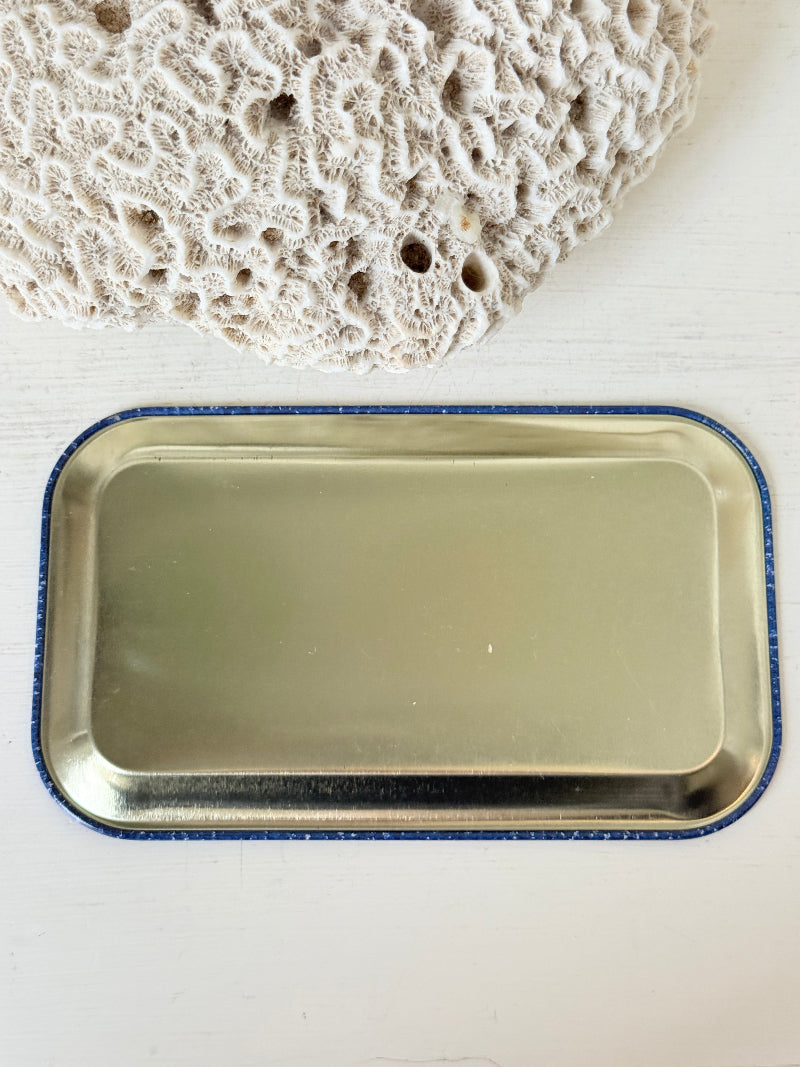 A rectangular metal tray with rounded corners and a blue glittery border, shown on a white surface with a textured beige stone prop in the upper corner.