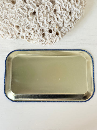 A rectangular metal tray with rounded corners and a blue glittery border, shown on a white surface with a textured beige stone prop in the upper corner.