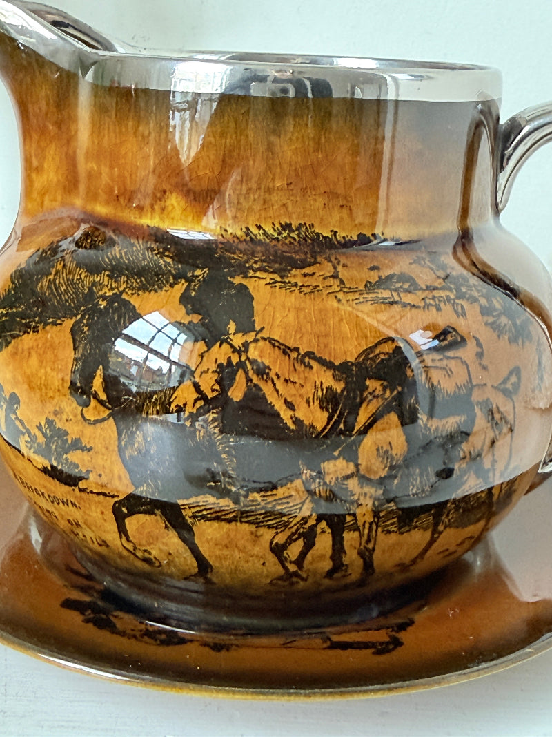 Antique Ridgways stoneware creamer jug and saucer photographed on a white surface in amber treacle glaze with black coaching scenes and silver lustre rim
