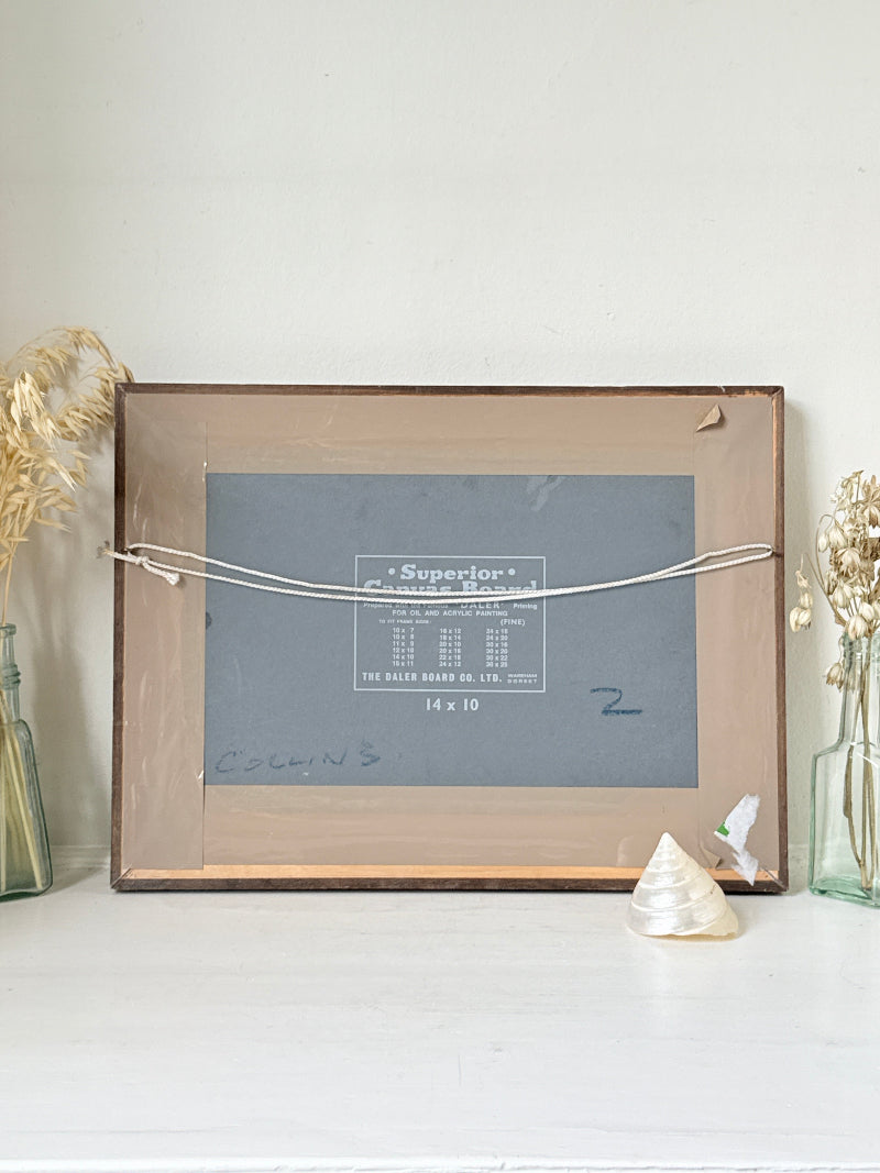 A vintage wooden picture frame with a clear glass front, held together by a white cord. The frame is set on a white surface against a white wall, flanked by dried grass in glass bottles and a small seashell.